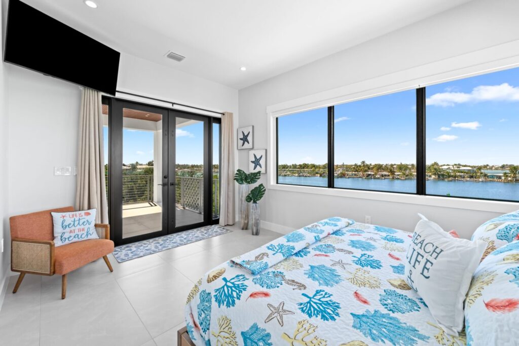Bright bedroom with coastal-themed decor, a large window overlooking water and palm trees, a bed with a colorful seashell quilt, a coral chair, wall-mounted tv, and sliding glass door to a balcony.
