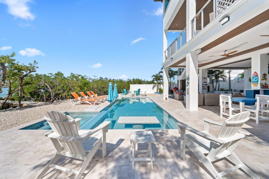 A luxurious outdoor pool area with white lounge chairs, sunbeds, and umbrellas, surrounded by tropical trees. The pool is next to a modern house with a covered patio, seating, and ceiling fans under a clear blue sky.