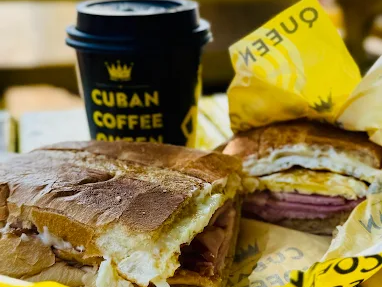 A cuban sandwich with ham, cheese, and egg sits on yellow branded paper next to a black takeaway cup labeled cuban coffee queen, all placed on a yellow wooden table.