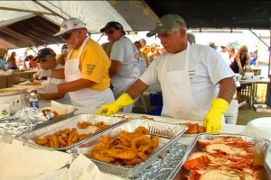 Several people in aprons and gloves serve fried food from large trays under a tent at an outdoor event, with a crowd of people visible in the background.