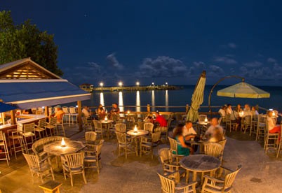 Outdoor restaurant by the waterfront at night, with groups of people sitting at wooden tables under warm lighting, and a pier illuminated in the background over calm water.