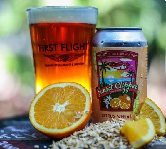 A can and pint glass of sunset clipper citrus wheat beer sit next to a sliced orange and wheat grains, with a blurred green background suggesting an outdoor setting.