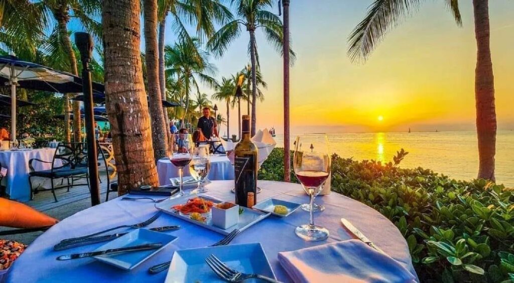 An outdoor restaurant table set for two with wine and appetizers overlooks the ocean at sunset. Palm trees, diners, and lush greenery surround the area under a colorful sky.