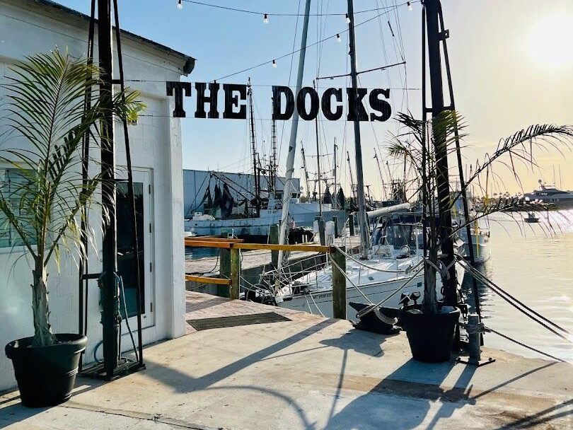 A dockside scene at sunset with boats moored in the water, palm trees in pots, and a sign reading the docks hanging between two poles near a white building.