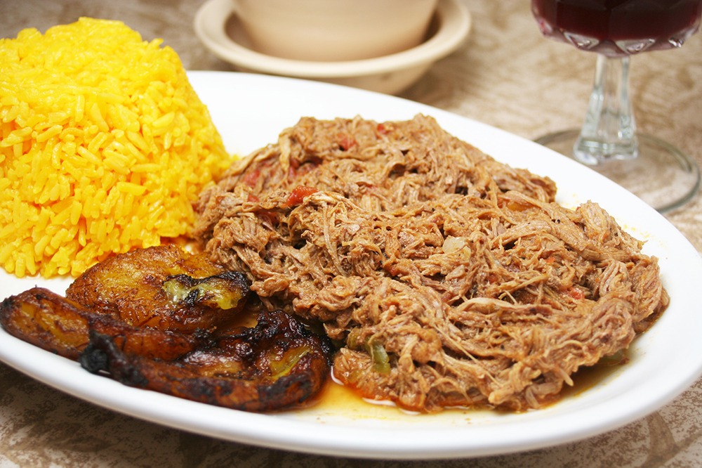A plate with yellow rice, shredded beef in sauce, and fried plantains, served on a white dish with a blurred glass and bowl in the background.