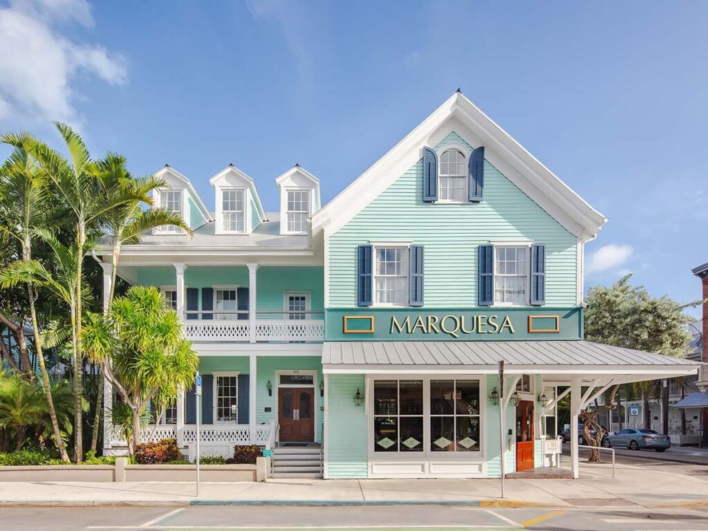 A light blue, two-story victorian-style building with white trim and three dormer windows, palm trees in front, and a sign that reads “marquesa” above the entrance on a sunny day.