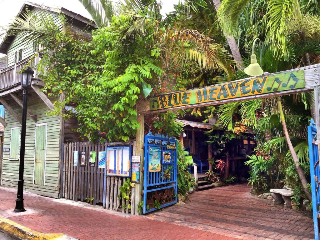 A vibrant, tropical restaurant entrance with a colorful “blue heaven” sign, lush green plants, wooden buildings, brick sidewalk, and blue gate, creating a welcoming, island atmosphere.