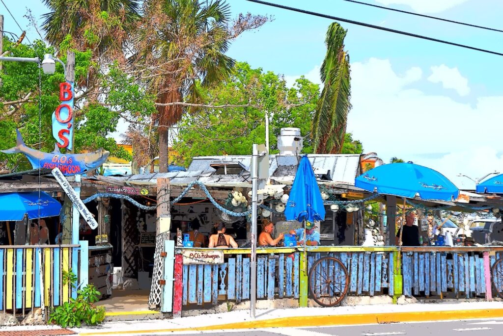 A colorful, open-air beach bar with blue umbrellas, rustic decor, and palm trees in the background. People are seated at tables, enjoying food and drinks in a relaxed, tropical setting under a bright sky.