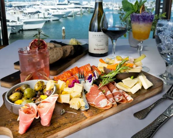 A wooden board with assorted cheeses, cured meats, olives, bread, and herbs sits on a table with wine, cocktails, and water, overlooking a marina with boats in the background.