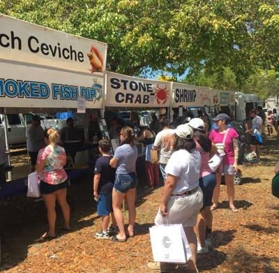 People stand in line at an outdoor food stall under trees, with signs advertising ceviche, smoked fish dip, stone crab, and shrimp. Many are holding bags and wearing casual clothes, enjoying a sunny day.
