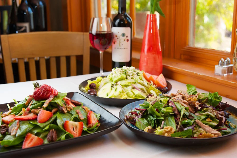 Three plates of fresh salads with mixed greens, strawberries, pecans, shredded meat, and cheese are on a table beside a glass and bottle of red wine, with a red vase and bright window in the background.