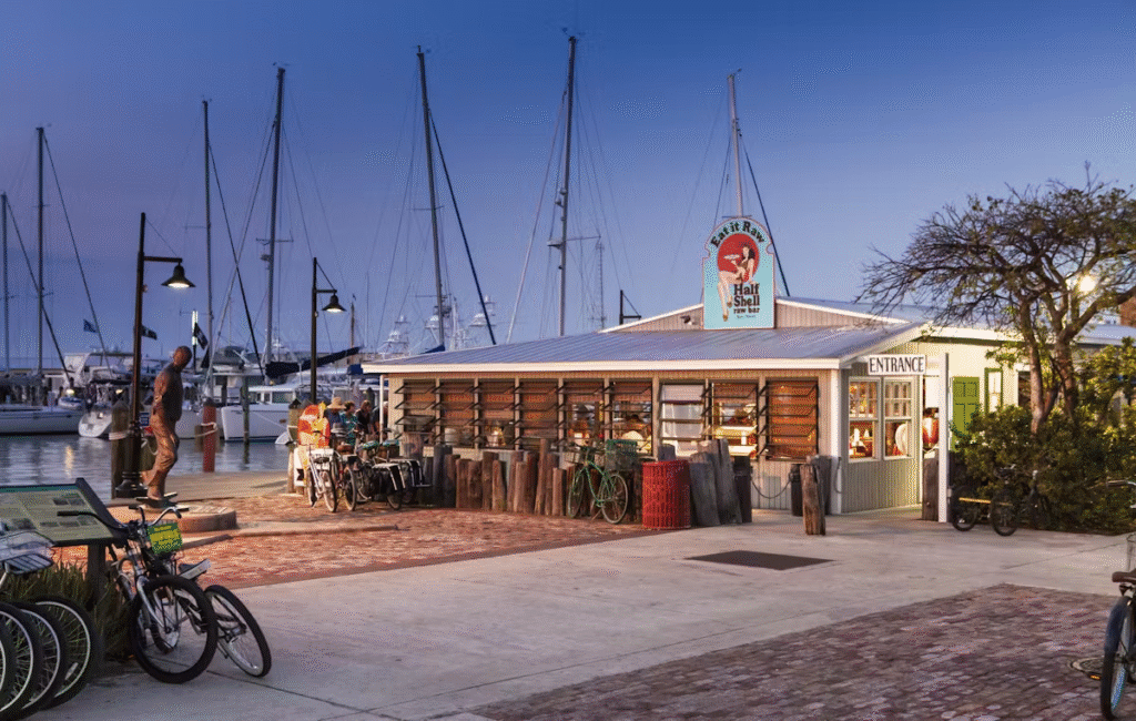 A seafood restaurant with a lobster sign sits by a marina with docked sailboats at dusk. Bicycles are parked outside, and people are gathered near the entrance under warm evening lights.