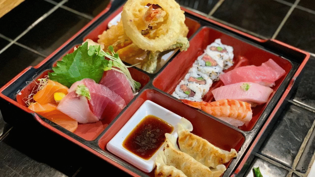 A bento box with assorted sushi, sashimi, tempura vegetables, gyoza dumplings, and a small dish of soy sauce, all arranged neatly on a black tiled surface.