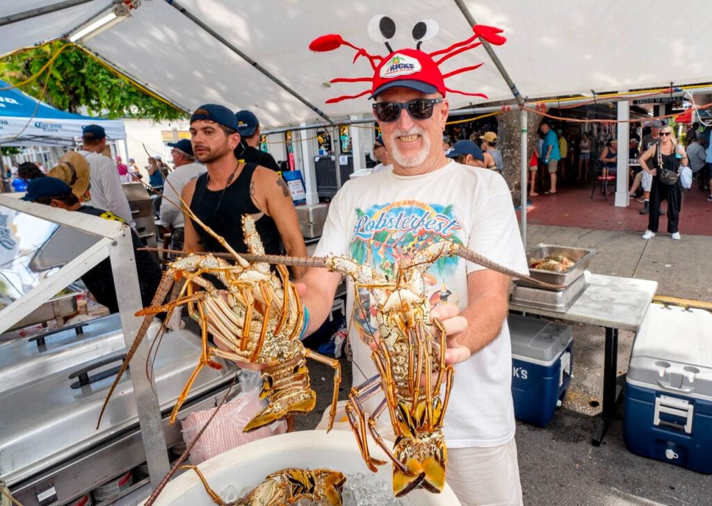 A smiling man wearing a red crab hat and sunglasses holds two large lobsters at an outdoor seafood festival booth, surrounded by coolers and other people under a white tent.