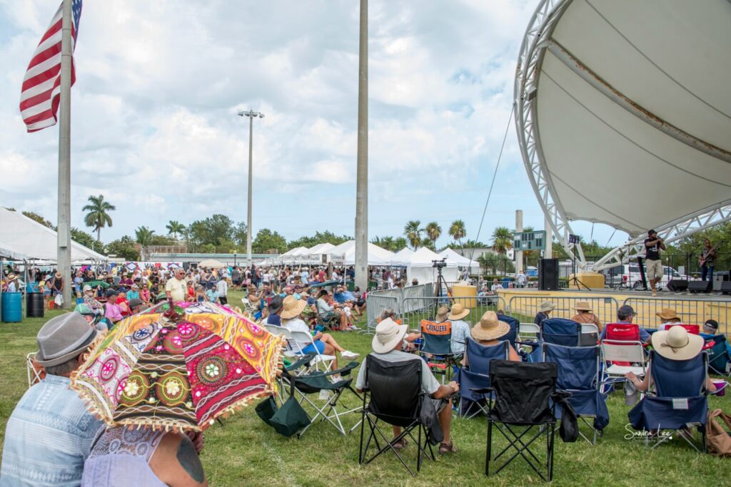 A crowd sits on lawn chairs facing an outdoor stage at a festival. An american flag flies on the left, and colorful umbrellas and hats dot the audience. Tents and palm trees are visible in the background under a partly cloudy sky.