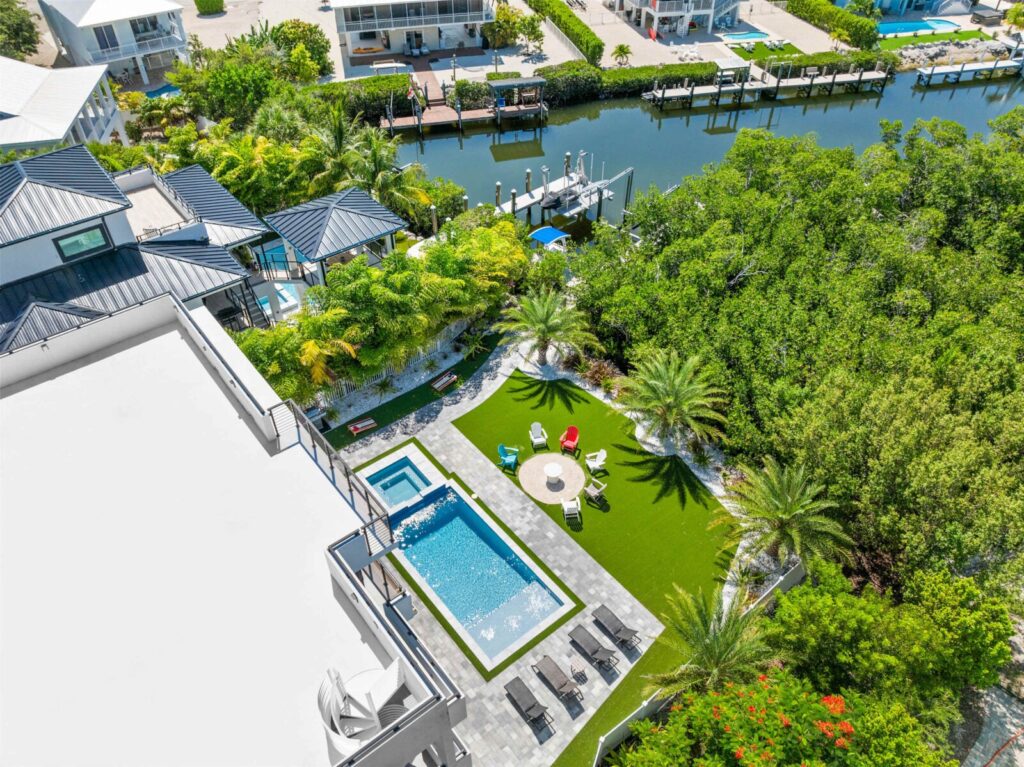 Aerial view of a modern backyard with a rectangular swimming pool, lounge chairs, a fire pit area, palm trees, and lush greenery near a canal with boat docks and waterfront homes.