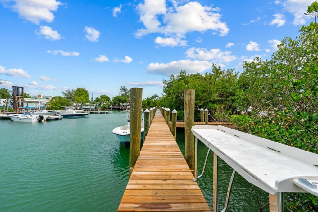 A wooden dock with rail posts stretches over calm blue-green water, leading to moored boats on both sides. Green trees border the right side, and the sky is bright with scattered clouds.