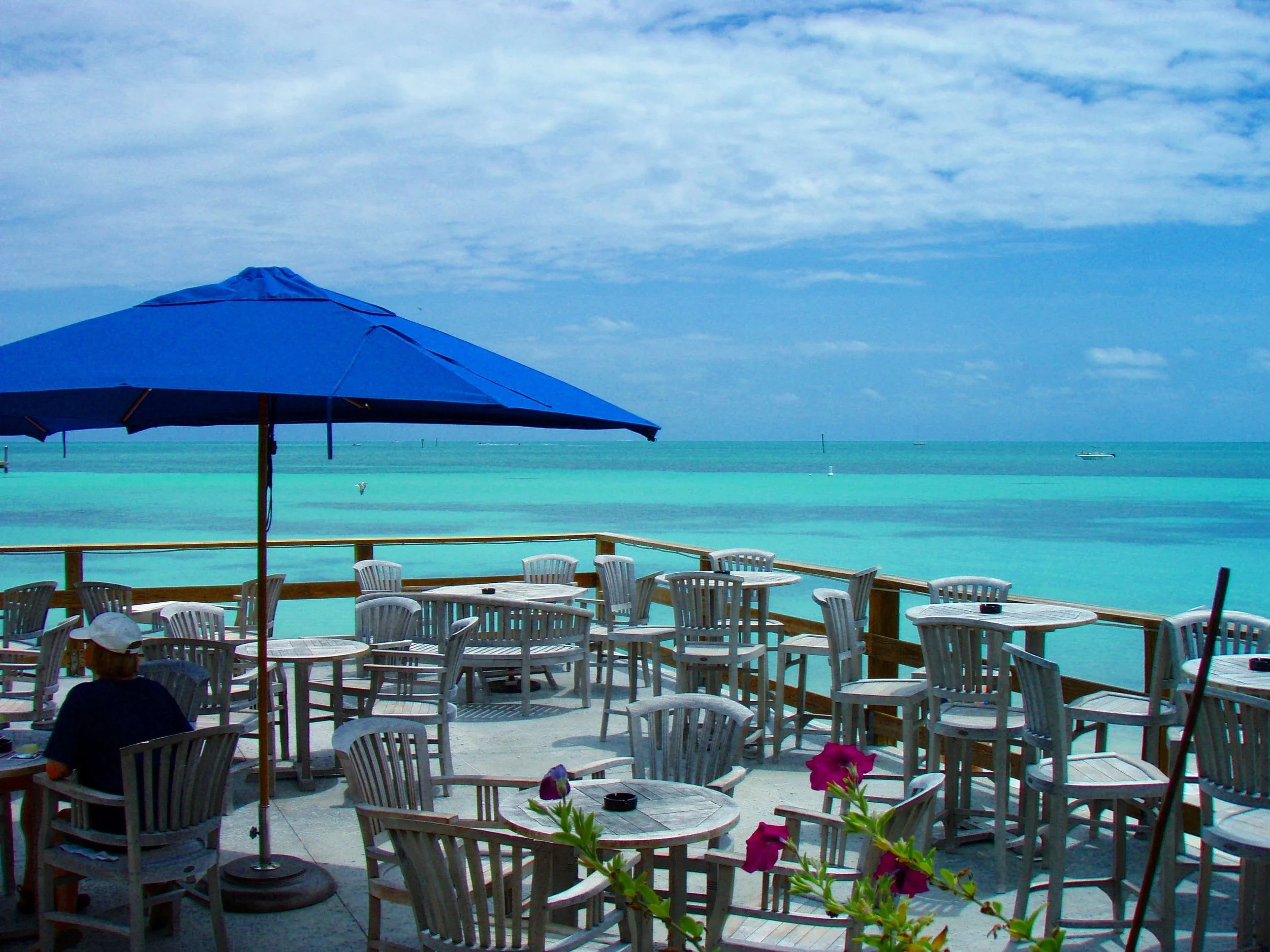 Outdoor patio with white tables and chairs overlooks a turquoise ocean under a partly cloudy sky. A blue umbrella provides shade, and a person sits alone. Pink flowers add charm, evoking the relaxed vibe of key west florida restaurants.