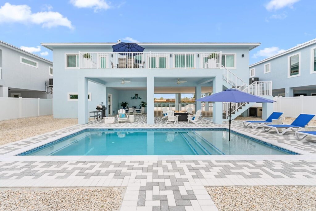 Modern two-story light blue house with an outdoor pool, lounge chairs, blue umbrellas, and a spacious patio area featuring seating and a grill, surrounded by white fencing and gravel landscaping.