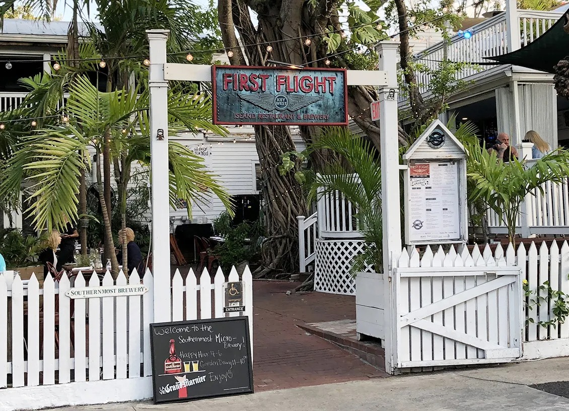 Entrance to first flight, one of the charming key west florida restaurants, features a white picket fence, chalkboard menu, and tropical plants. A two-story white building with outdoor seating is visible in the background.