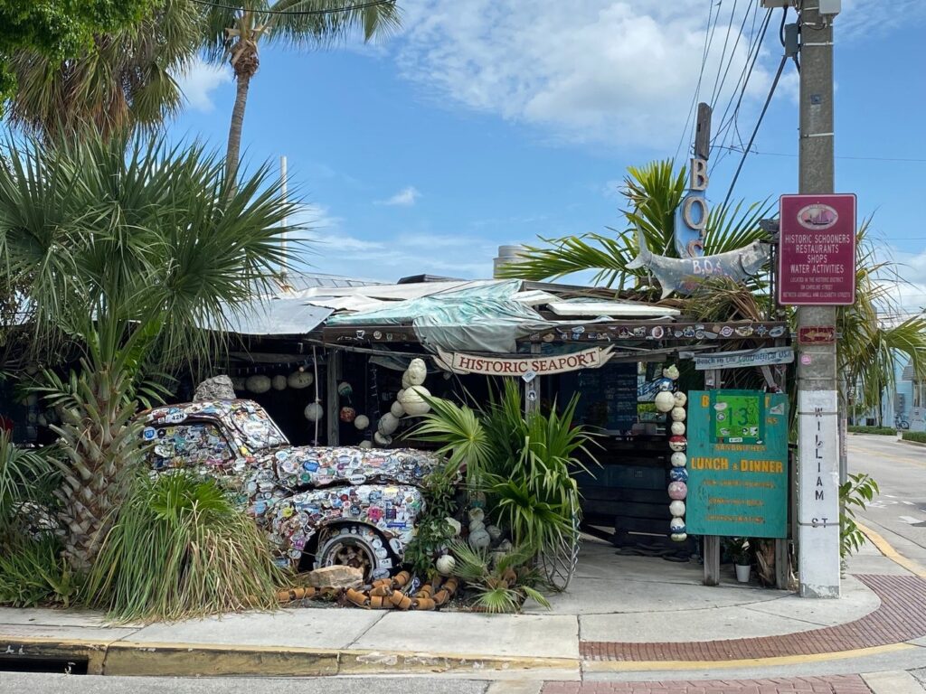 A rustic, weathered shack adorned with tropical plants and buoy decorations sits at a corner. A classic truck, covered in stickers, is parked partly outside. Signs reveal it's part of the historic seaport and one of the unique key west florida restaurants serving lunch and dinner.