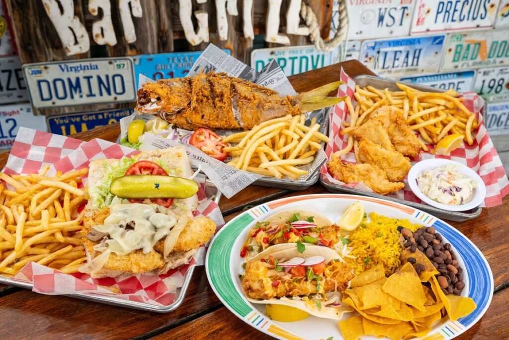 A table filled with fried fish, fries, a sandwich, coleslaw, fish tacos with chips, rice, black beans, and garnishes, set in front of a wall decorated with license plates.