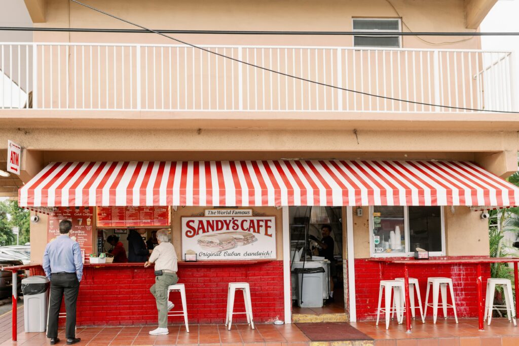 A classic spot among key west florida restaurants, this cafe features a red-and-white striped awning, a red brick facade, and a sign reading sandys cafe, original cuban sandwich. Several people gather outside by stools and a counter on the ground floor.