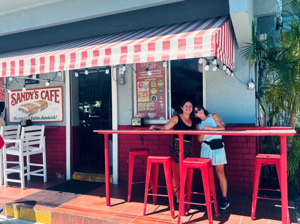 Two women smile and hug while standing at a red outdoor table in front of sandy’s cafe, which has a red-and-white striped awning and menu signs on the wall.