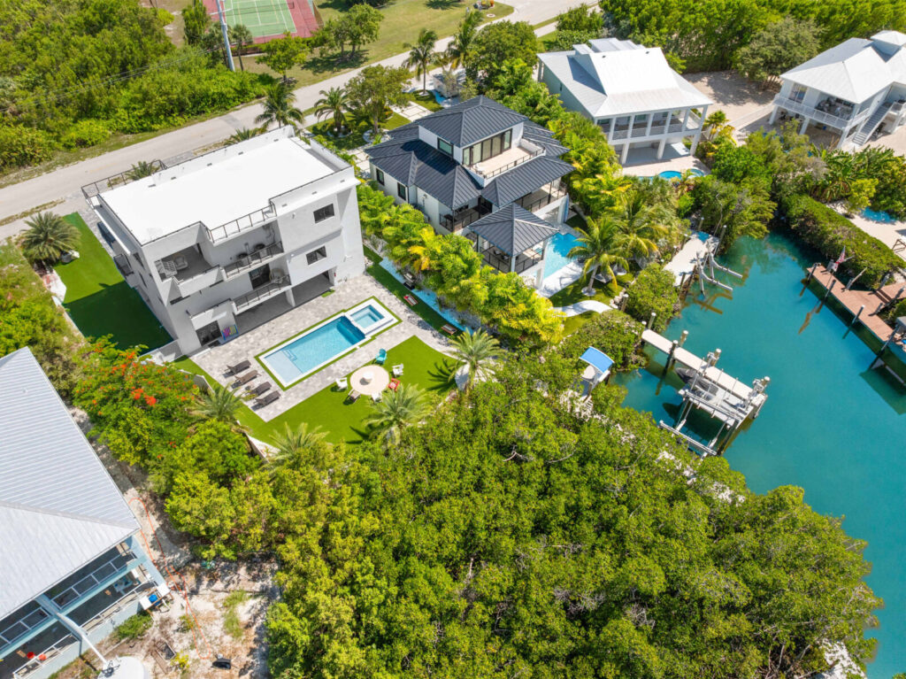 Aerial view of a modern house with a pool and lawn, surrounded by palm trees, next to a waterway with docks and boats; other houses and a tennis court are visible nearby.