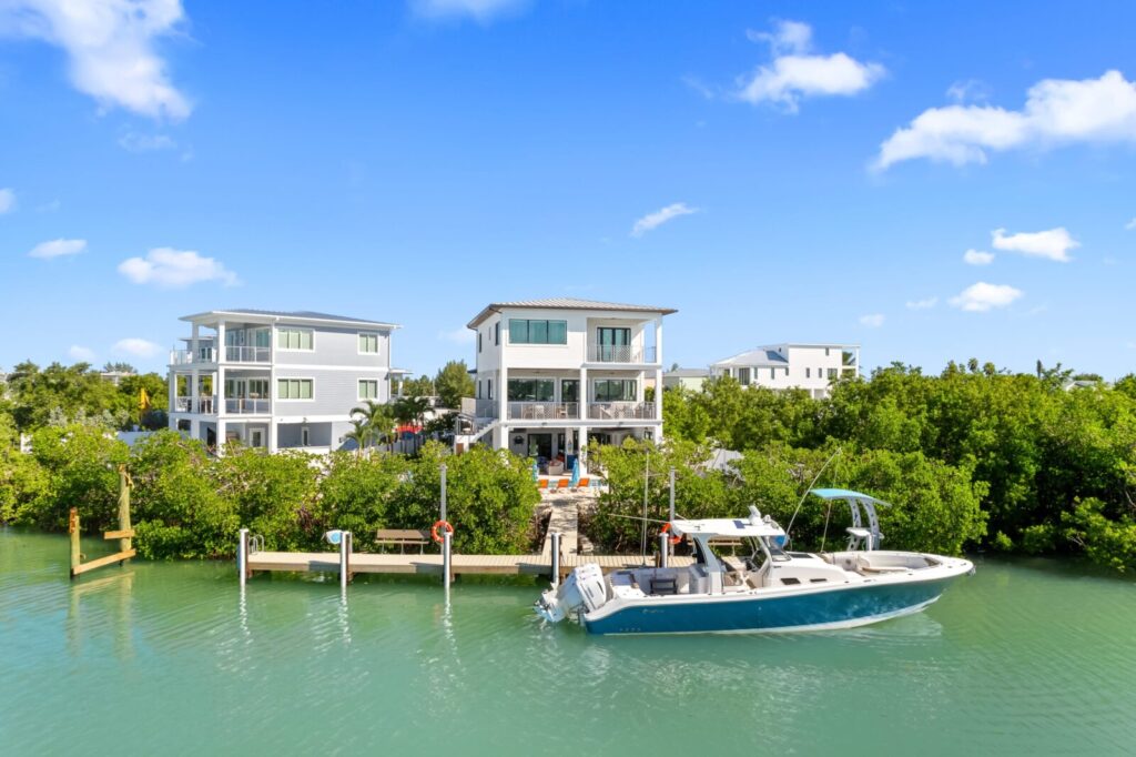 A modern waterfront house with large windows sits behind lush greenery and a docked speedboat on a calm, turquoise canal, under a bright blue sky with scattered clouds.