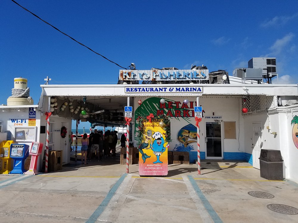 A sunny exterior view of a colorful seafood restaurant and marina with a cartoon fish sign, festive decorations, and people entering through an open doorway under a blue sky—perfect for any marathon fl seafood festival guide.