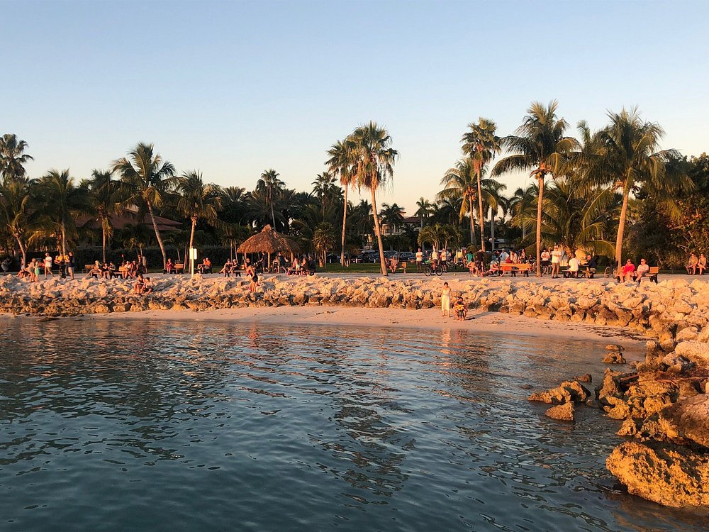 A sandy beach lined with palm trees at sunset, with many people relaxing and walking along the shore. The calm water reflects the warm light, and rocks border parts of the shoreline.