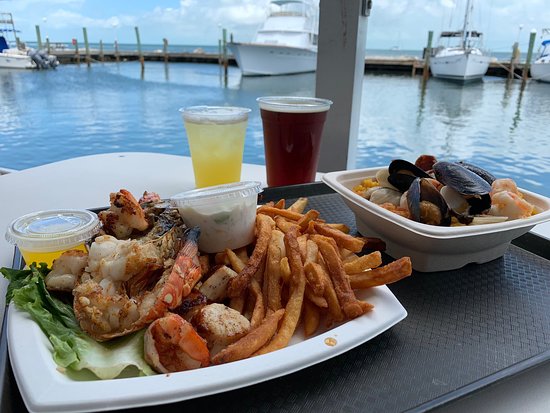 A plate of grilled seafood, fries, and lemon sits on a table next to a bowl of seafood stew and two drinks, overlooking a marina with boats docked under a partly cloudy sky.
