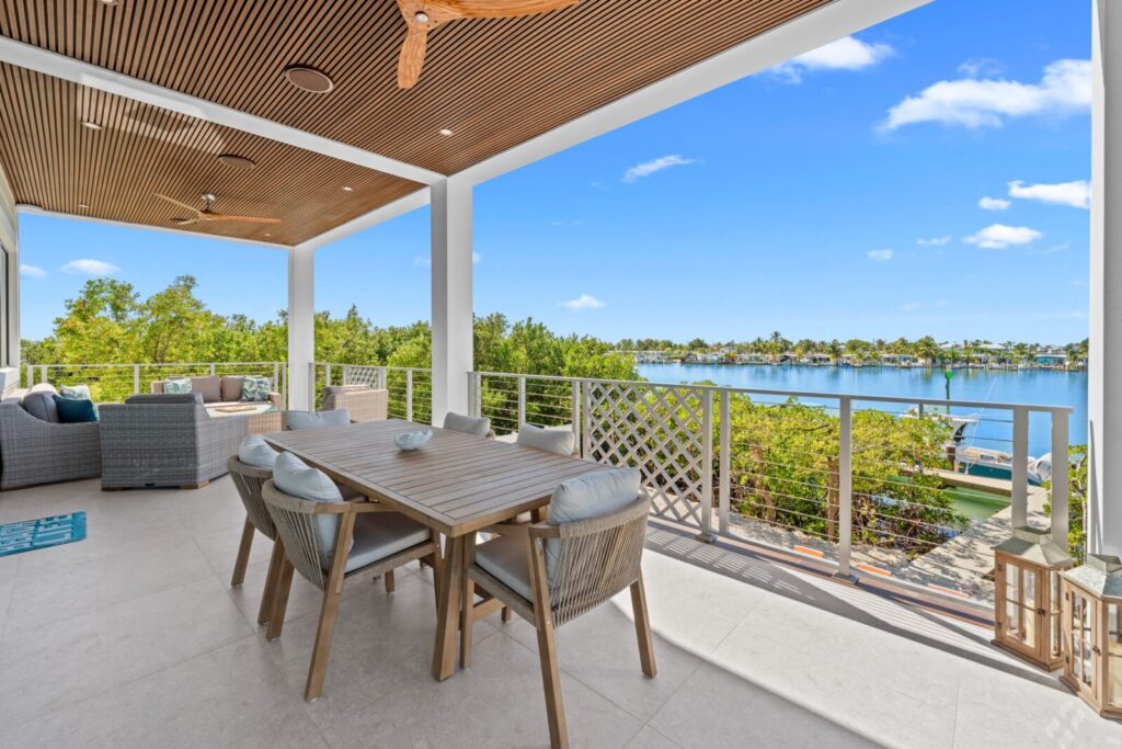 A spacious outdoor patio with a wooden dining table and chairs overlooks a peaceful lake. Lounge seating is in the background, and the patio is shaded by a wood-paneled ceiling with fans. Lush greenery surrounds the area.