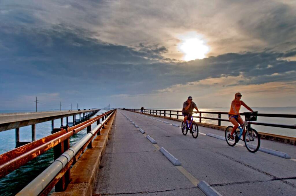 Two people ride bicycles on a wide bridge over water at sunset, with dramatic clouds in the sky and metal railings on both sides of the bridge.