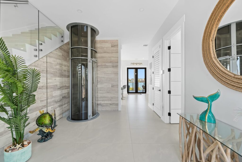 Modern home entryway with light tile floors, wooden accent walls, a glass tube elevator, and coastal decor including a plant, a globe, and a mermaid sculpture. Glass doors at the back reveal water views.