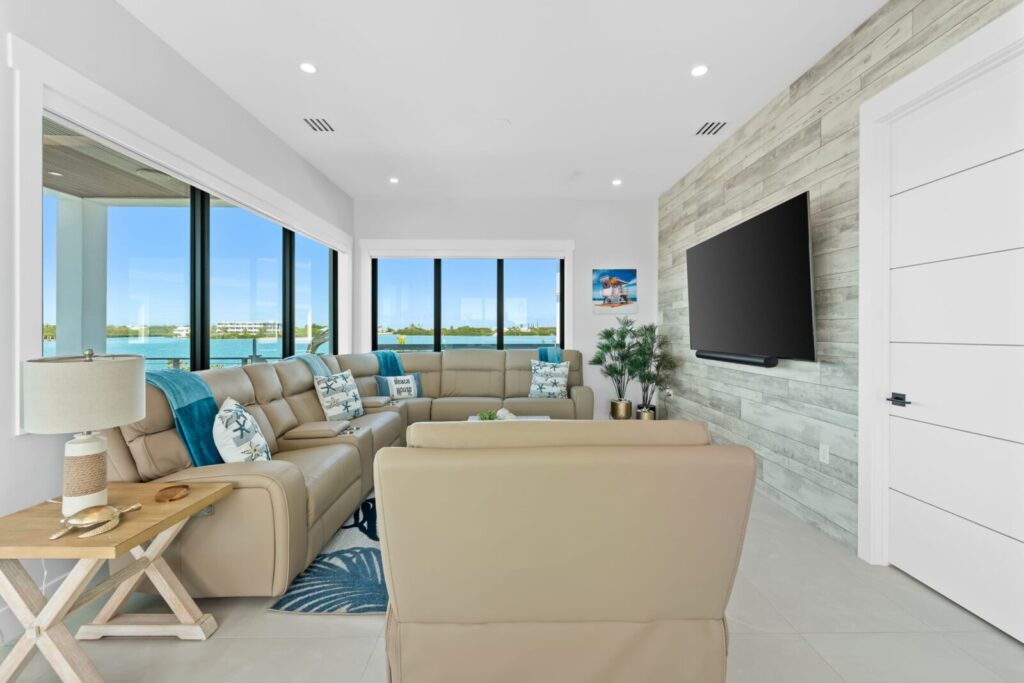 Modern living room with beige leather sofas, coastal-themed pillows, a wooden accent wall with a large tv, a side table with a lamp, and large windows offering a scenic water view.