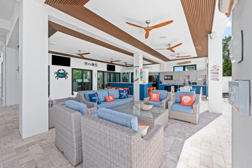 Spacious outdoor patio with wicker seating, blue cushions, and orange accent pillows. Ceiling fans, wall decor, and a modern kitchen with blue cabinets are visible. The area is bright and airy with natural light.