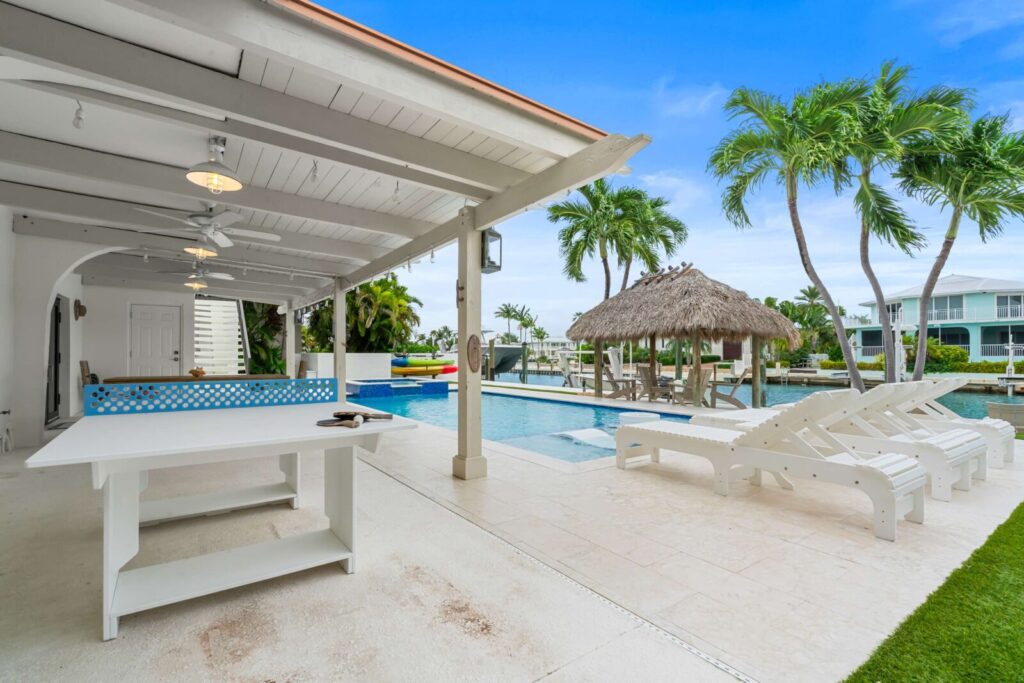 Covered patio with a white ping-pong table, paddle and ball, lounge chairs, a thatched tiki hut, and a pool surrounded by palm trees, with modern houses in the background under a blue sky.