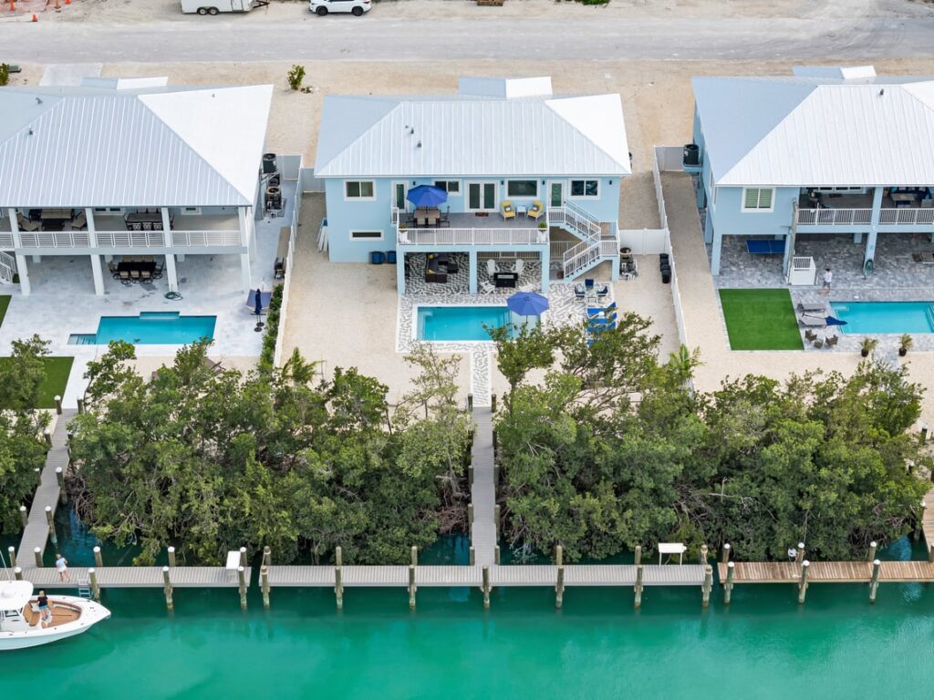 Aerial view of waterfront houses with pools and docks, separated by trees from turquoise canal water; a boat is docked near one house, and cars are parked on the street behind the homes.
