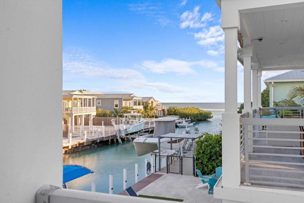 View from a covered porch overlooking a canal lined with modern houses, boats docked along the water, and a glimpse of the ocean horizon under a blue sky with scattered clouds.