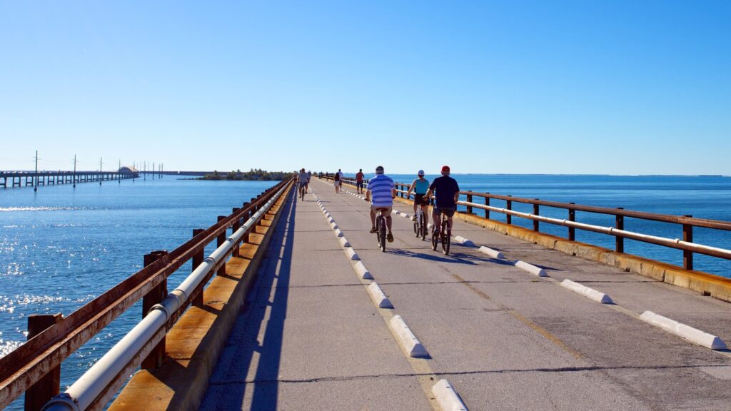People ride bicycles on a long, narrow bridge over blue water under a clear sky. The bridge has railings on both sides and stretches into the distance.
