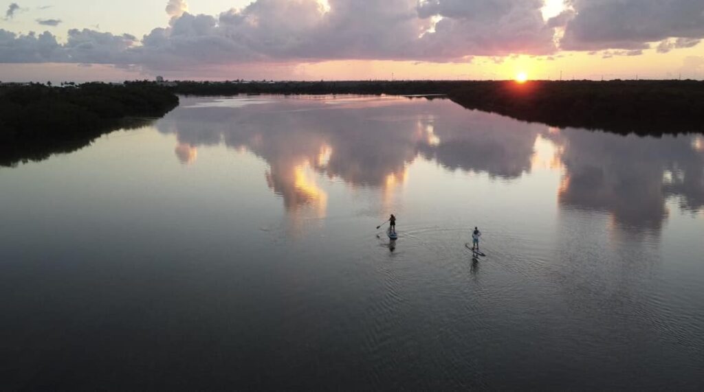 Two people paddleboarding on a calm river at sunset, with clouds and the sun reflected on the water and trees lining the horizon.