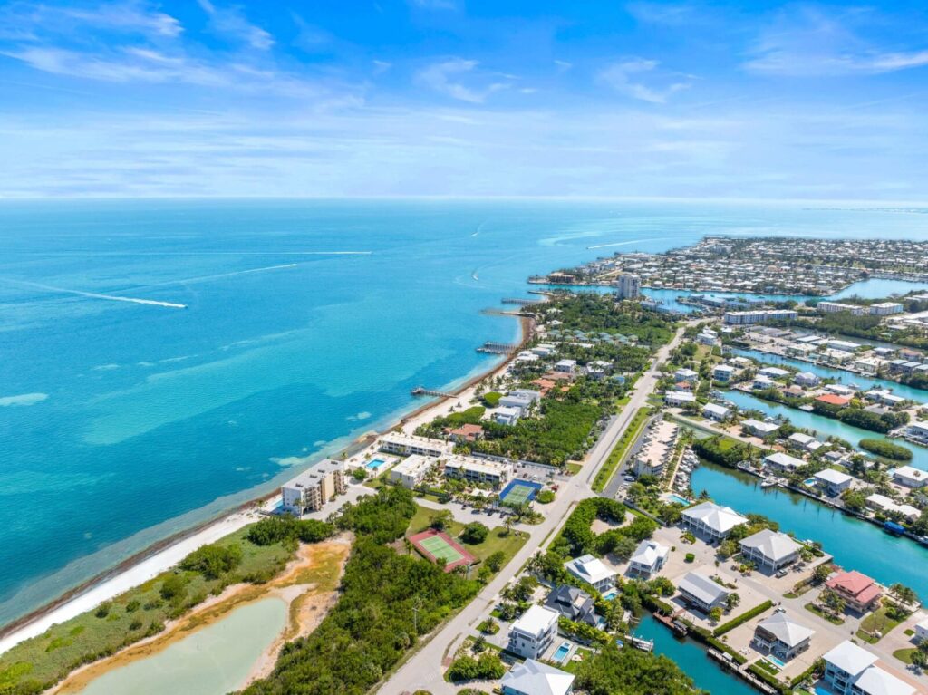 Aerial view of a coastal town with turquoise ocean, sandy shoreline, green trees, residential houses, and winding roads under a bright blue sky.
