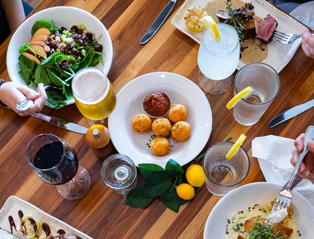 Dining in florida keys: a table set with assorted dishes, including a spinach salad, fried cheese balls with dip, steak, pasta, and drinks like beer, red wine, and water with lemon. Several hands are reaching for food and eating.