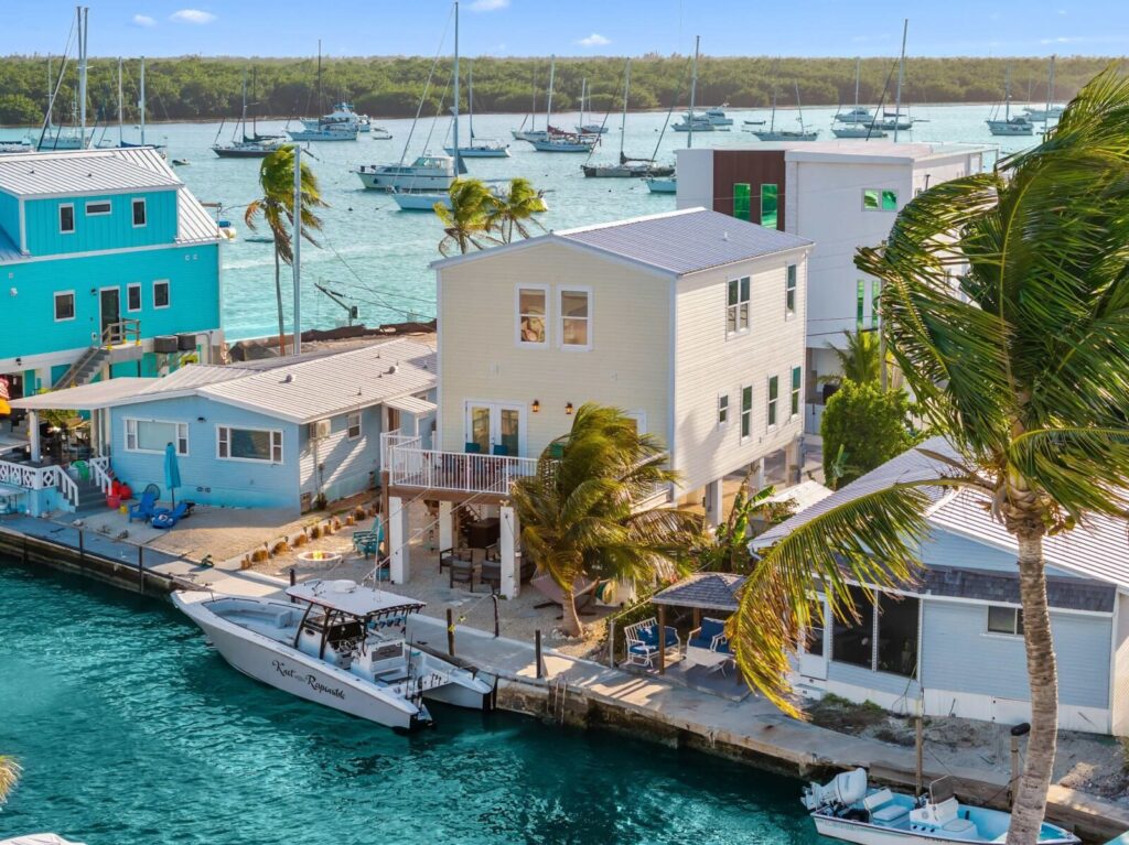 A white two-story house by a turquoise canal with a docked boat, surrounded by palm trees and colorful houses; sailboats float in the background on calm water.