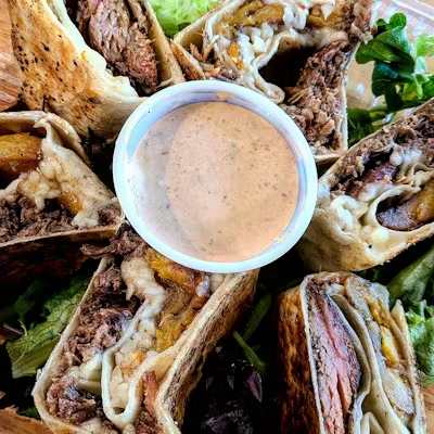 Sliced burritos filled with beef, melted cheese, and plantains arranged around a cup of creamy dipping sauce, served on a bed of fresh greens.