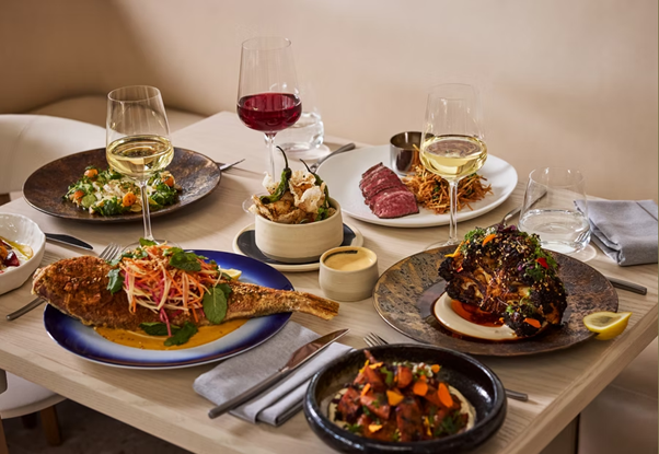 A table set with various gourmet dishes, including a whole fried fish, grilled meat, vegetables, sauces, and three glasses of wine, arranged neatly on stylish plates in a modern dining setting.