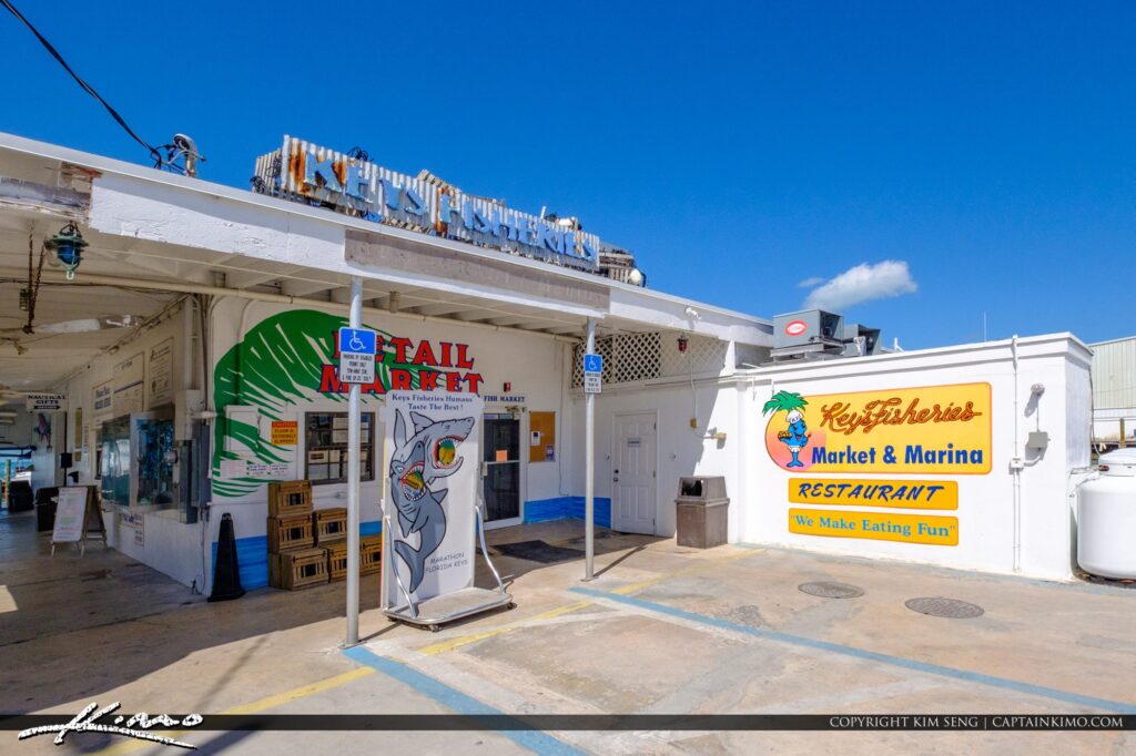 A sunny exterior view of key fisheries market & marina restaurant, featuring colorful signs, a fish mural, and a shaded entryway with palm trees and blue sky above.
