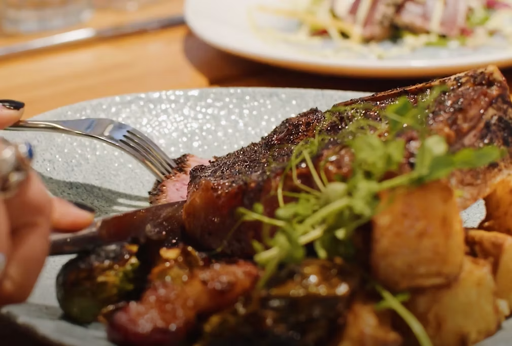 A close-up of a plate with a grilled steak garnished with greens, roasted vegetables, and potatoes. A hand is cutting into the steak with a fork and knife. Another dish is visible blurred in the background.